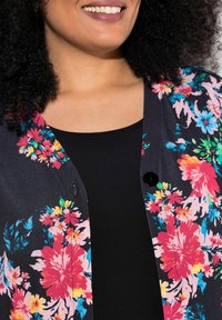 Bloemen cardigan met een zwarte achtergrond, versierd met felroze, blauwe en gele bloemen, knoopsluiting en een zachte textuur.