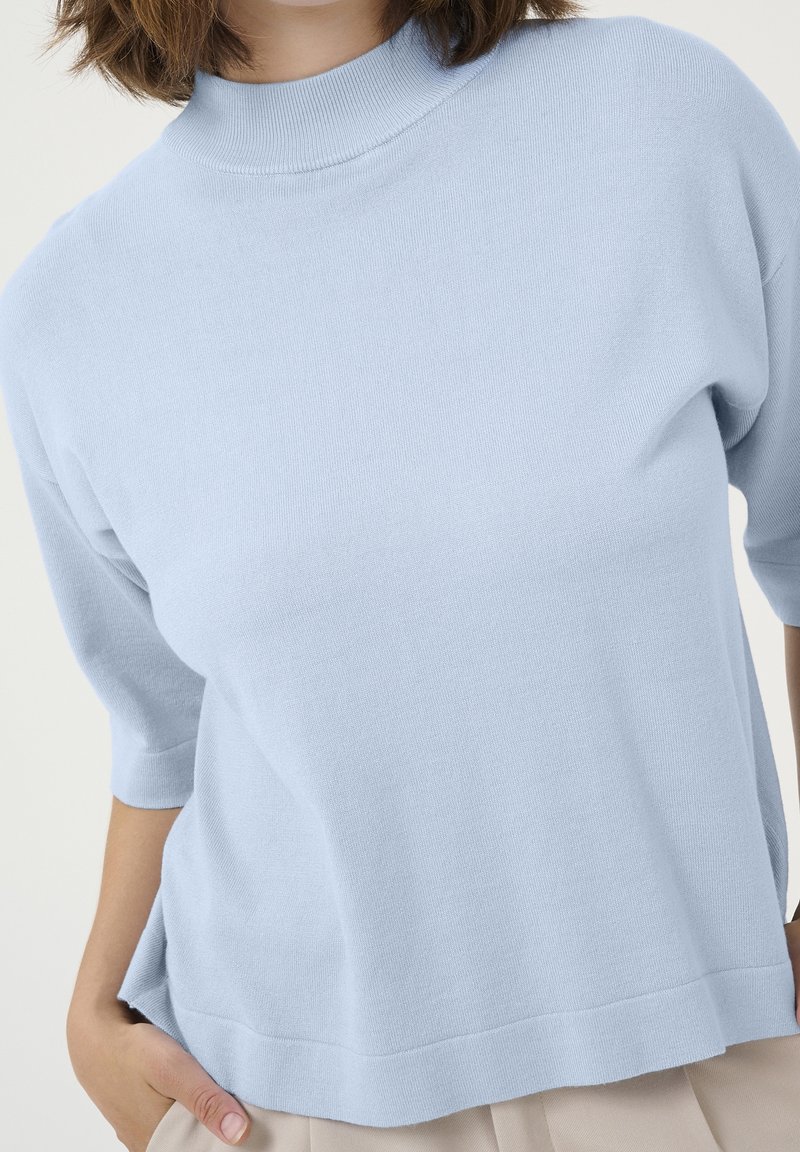Ljusblå stickad topp med hög rundhals och korta ärmar. Har en något löst sittande passform och en slät yta.