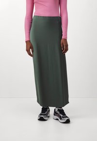Lange olijfgroene rok met een gladde textuur, gecombineerd met een aansluitend roze langslavend top en zwarte sportieve schoenen met zilveren accenten.