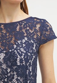 Žena v námořnické modré květinové krajkové blůze s krátkými rukávy a kulatým výstřihem, pod kterou má světlý oděv, na jednolitém pozadí.