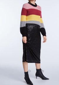 Una donna indossa un maglione a maglia a righe con fasce rosa, bordeaux, giallo, grigio e nero, una gonna midi nera con paillettes e stivali neri con lacci alla caviglia.