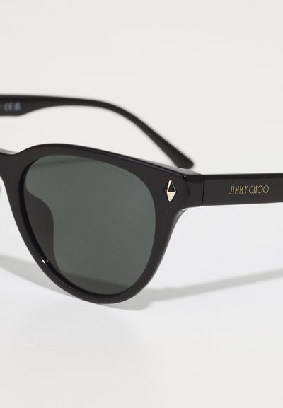 Černé plastové sluneční brýle s šedými čočkami, kulatým rámem a zlatým akcentem na spánku s nápisem značky "JIMMY CHOO."