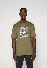 Mand iført en olivengrøn camo-print grafisk T-shirt med hvid tekst og sorte bukser, stående mod en ensfarvet lys baggrund.
