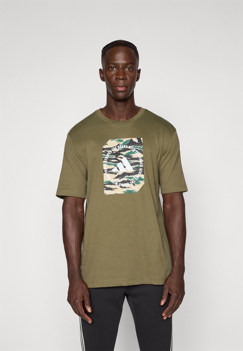 Mand iført en olivengrøn camo-print grafisk T-shirt med hvid tekst og sorte bukser, stående mod en ensfarvet lys baggrund.