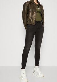 Camisa bomber de cuero verde oliva, con puños acanalados, jeans ajustados de cintura alta negros, zapatillas gruesas blancas con detalles reflectantes.