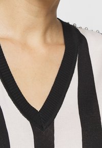 Czarny i biały sweter w paski z dekoltem w serek, z prążkowanymi wykończeniami i ozdobnymi perłami wzdłuż szwów na ramionach. Miękka tekstura.