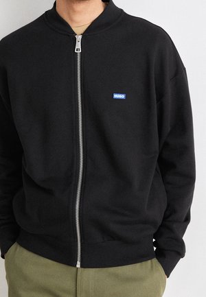 Man wearing black zip-up jacket with small blue "HUGO" logo on the chest and olive green pants, hands in pockets.