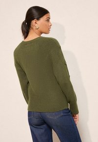 Pulover verde măslinie cu un model de tricotaj texturat, tiv ribat, și accente cu nasturi pe mâneci, asortat cu blugi negri.