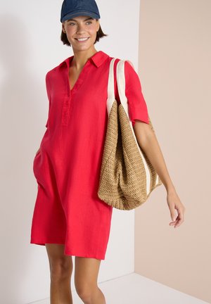 Femme souriante portant une robe rouge en lin à manches courtes et une casquette bleue, portant un grand sac cabas beige tissé sur son épaule.