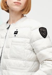 Personne portant une veste matelassée blanche avec une poche frontale, une étiquette Blauer U.S.A. et un écusson noir brodé sur la manche supérieure.