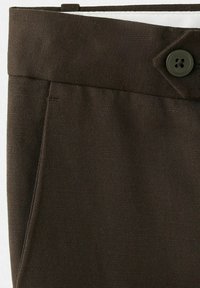 Mørkebrune, teksturerede bukser af stof med en talje med knap og bæltestrop, foldet sammen mod en hvid baggrund.