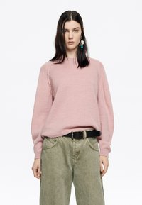Maglione a maglia rosa chiaro con maniche lunghe e svasate e scollatura rotonda, abbinato a pantaloni a gamba larga verde oliva e una cintura nera.