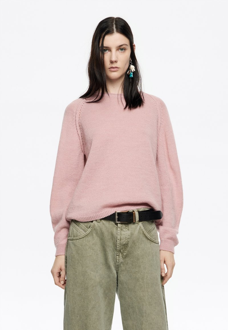 Maglione a maglia rosa chiaro con maniche lunghe e svasate e scollatura rotonda, abbinato a pantaloni a gamba larga verde oliva e una cintura nera.
