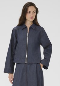 Denim zip-up jacket, dark blue with brown stitching, collar, and side pockets; relaxed fit with wide sleeves. Smooth texture.