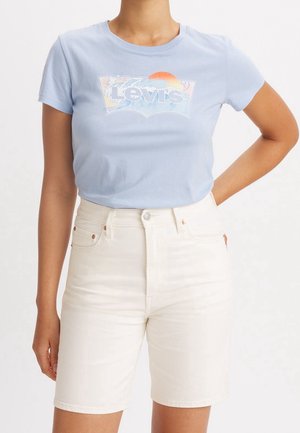 Persona vistiendo una camiseta Levi's de color azul claro con un gráfico de ola y sol, combinada con pantalones cortos de denim blanco de cintura alta, de pie con una mano en la cadera.