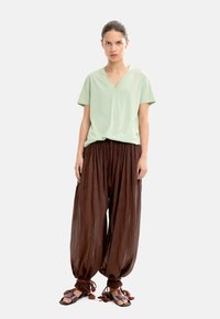 Jasnozielona koszulka V-neck, luźne brązowe spodnie haremowe i czarne sandały z czerwonymi sznurkami i frędzlami. Gładki materiał o płynnej fakturze.