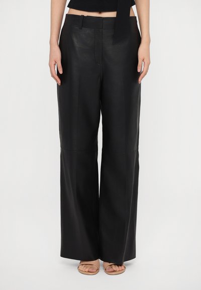 Pantalons larges en cuir noir avec une texture lisse, présentant une taille cintrée et des coutures discrètes à l'avant, associés à des sandales beiges.