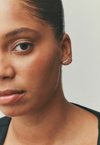 Sølvørering med et buet design, med tre små klare ædelstene. Nærbillede af en kvinde, der viser øreringen på sit venstre øre.