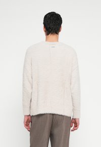 Bărbat cu păr scurt și închis la culoare, purtând un pulover pufos de culoare alb-crem și pantaloni maro cu dungi, stând cu fața spre un perete simplu.