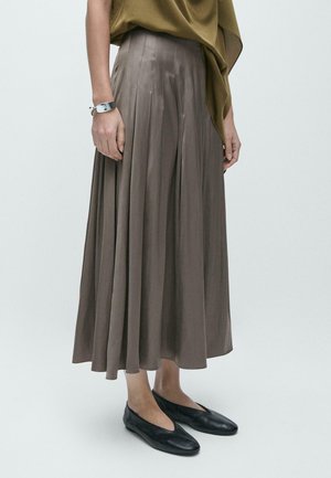 Vrouw die een taupekleurige geplooide midirok, een olijfgroen topje, een zilveren armband en zwarte platte schoenen draagt, staand tegen een effen lichte achtergrond.