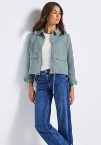 Ljusgrön jacka med dragkedja fram, krage, två framfickor och justerbar dragsko i nederkant, kombinerad med blåjeans.