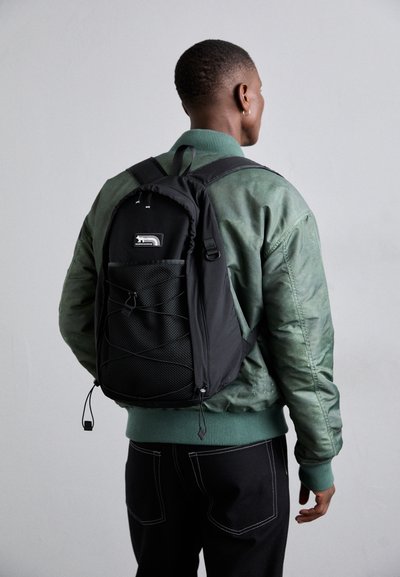 Un sac à dos noir avec une poche frontale en filet, des bretelles ajustables et des détails en cordon élastique, porté sur une veste bomber verte et un pantalon noir.