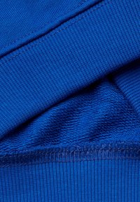 Tessuto blu con una finitura testurizzata, caratterizzato da polsini a costine e sezioni lisce. Il materiale appare morbido e leggermente peloso.