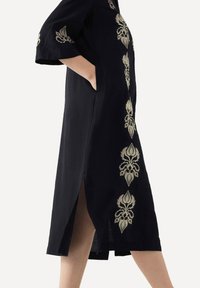 Schwarzes Leinenkleid mit goldenen Blumenstickereien an den Seiten, kurzen Ärmeln und seitlichen Schlitzen für eine bequeme Bewegungsfreiheit.