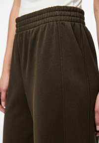 Pantalons de jogging marron avec une large ceinture élastique, présentant des coutures verticales et un tissu doux et texturé. Gros plan de la taille et du côté.