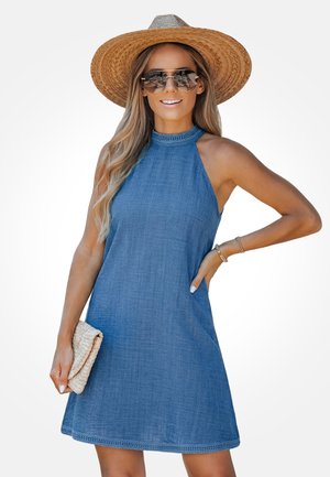 Vrouw draagt een mouwloze blauwe jurk, grote strohoed, zonnebril en armbanden, met in één hand een beige gevlochten clutch.