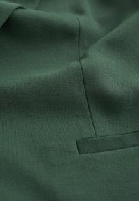 Tessuto verde con una trama testurizzata, caratterizzato da una leggera lucentezza. È visibile una piccola tasca, integrata nel design del capo.