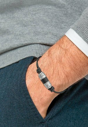 Braccialetto in gomma nera con accenti metallici argento, dotato di un pezzo centrale cilindrico, indossato su un polso con peli chiari, sopra un tessuto blu.