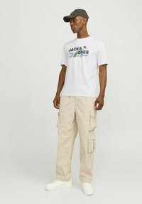 Camiseta blanca con texto gráfico azul y verde, pantalones cargo beige con múltiples bolsillos y zapatillas blancas. Gorra oscura en la cabeza.