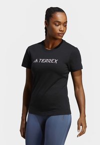 Γυναίκα φορώντας μαύρο T-shirt Adidas Terrex και μπλε κολάν, στέκεται με το κεφάλι στραμμένο ελαφρώς στο πλάι μπροστά σε απλό φόντο.