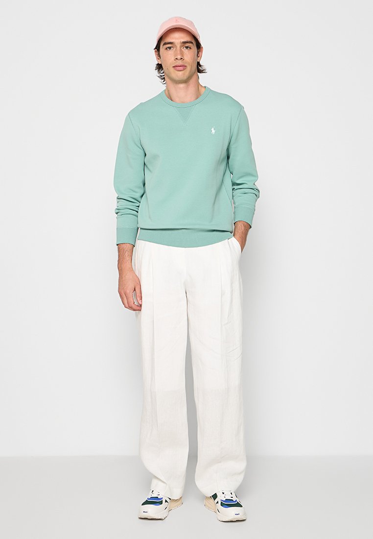 Mann in mintgrünem Sweatshirt, weißen weit geschnittenen Hosen, pinker Kappe und weißen Sneakers, der vor einem einfarbigen Hintergrund steht.