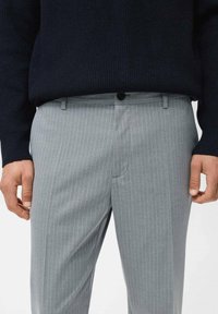 Pantalones a rayas gris con tejido suave y corte ajustado, que cuentan con un cierre frontal de botón y bolsillos laterales. Llevados con un suéter oscuro.