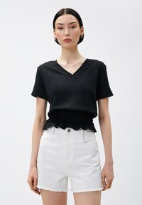 Sort kortærmet top med V-udskæring og tekstureret stof; samlet smocked taljedetalje. Parret med hvide højtaljede denimshorts.