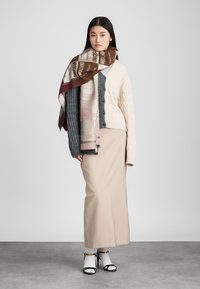 Bežinė ilga sijonas, kreminė pynimo megztinis, pilkas šalikas su tekstiniu raštu ir juodai balti sandalai su dirželiais. Lygūs tekstilės paviršiai, sluoksnių išvaizda.