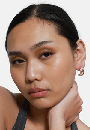 Orecchino a cerchio in metallo color oro rosa con una finitura liscia e lucida, che si abbina a un modello con un trucco naturale e lunghi capelli scuri.
