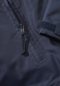 Lähikuva tummansiniseen takkiin, jossa on osittain auki oleva vetoketju kangaspidikkeellä ja hihansuu tarrakiinnityksellä.