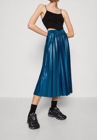 Gonna midi plissettata metallizzata turchese con vita aderente, abbinata a un top corto nero e sneakers sportive nere. Texture liscia nel complesso.