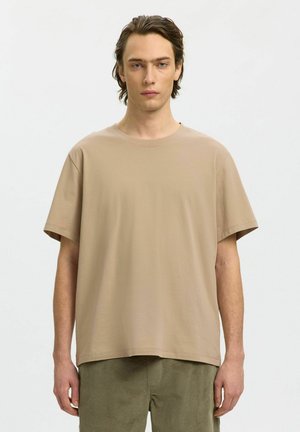 Jeune homme aux cheveux bruns mi-longs, portant un t-shirt beige ample à manches courtes et un pantalon vert olive, debout devant un fond uni.