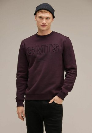 Felpa viola scuro con il testo grafico nero "GMTS" e scollatura rotonda. Presenta maniche lunghe e polsini e orlo a costine. Indossata con pantaloni neri.