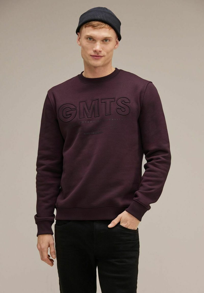 Felpa viola scuro con il testo grafico nero "GMTS" e scollatura rotonda. Presenta maniche lunghe e polsini e orlo a costine. Indossata con pantaloni neri.