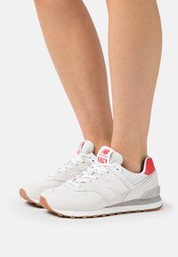 Pair of white and red New Balance 574 sneakers worn on feet, featuring mesh and suede uppers with gum soles on a white background.