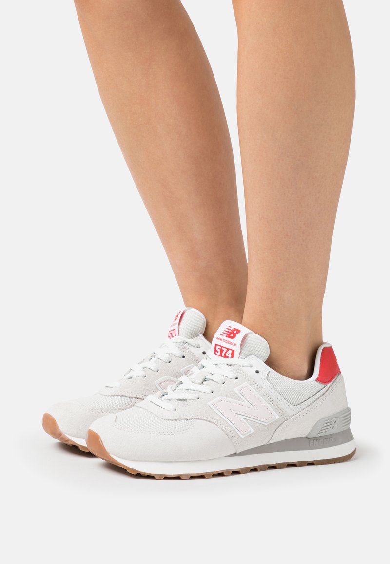 Pair of white and red New Balance 574 sneakers worn on feet, featuring mesh and suede uppers with gum soles on a white background.