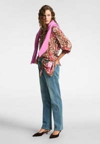 Donna che indossa una blusa a motivo marrone e rosa, jeans blu con una sciarpa annodata, tacchi scuri a scarpa a sella e un maglione rosa gettato sulle spalle.
