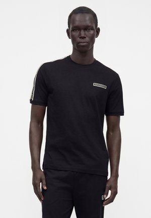 Homme portant un t-shirt noir à manches courtes avec un logo blanc sur la poitrine et un détail à rayures sur les manches, associé à un pantalon noir sur un fond uni.