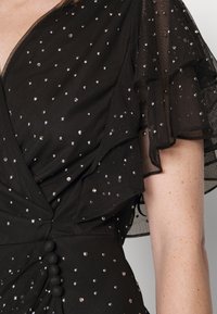 Svart klänning med volangärmar i genomskinligt material, prydd med små rhinestones. Har en insvängd design och dekorativa knappar längs midjan.