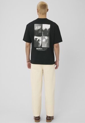 Personne aux cheveux blonds courts, vue de dos, portant un t-shirt noir avec quatre impressions photo noir et blanc florales et abstraites dans le dos.